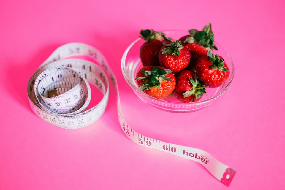 Cinta métrica junto a un tazón de fresas sobre fondo rosa