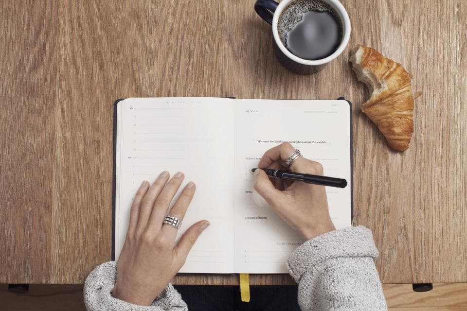 Persona escribiendo en una libreta junto a un café y un cuernito