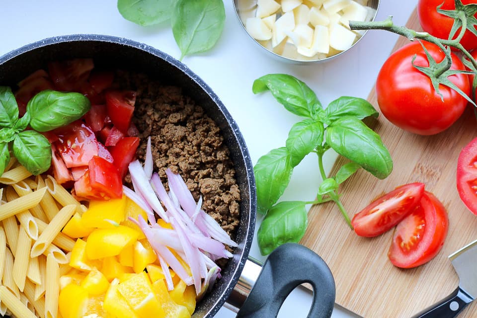 Pan with pasta, meat and vegetables next to tomatoes and basil